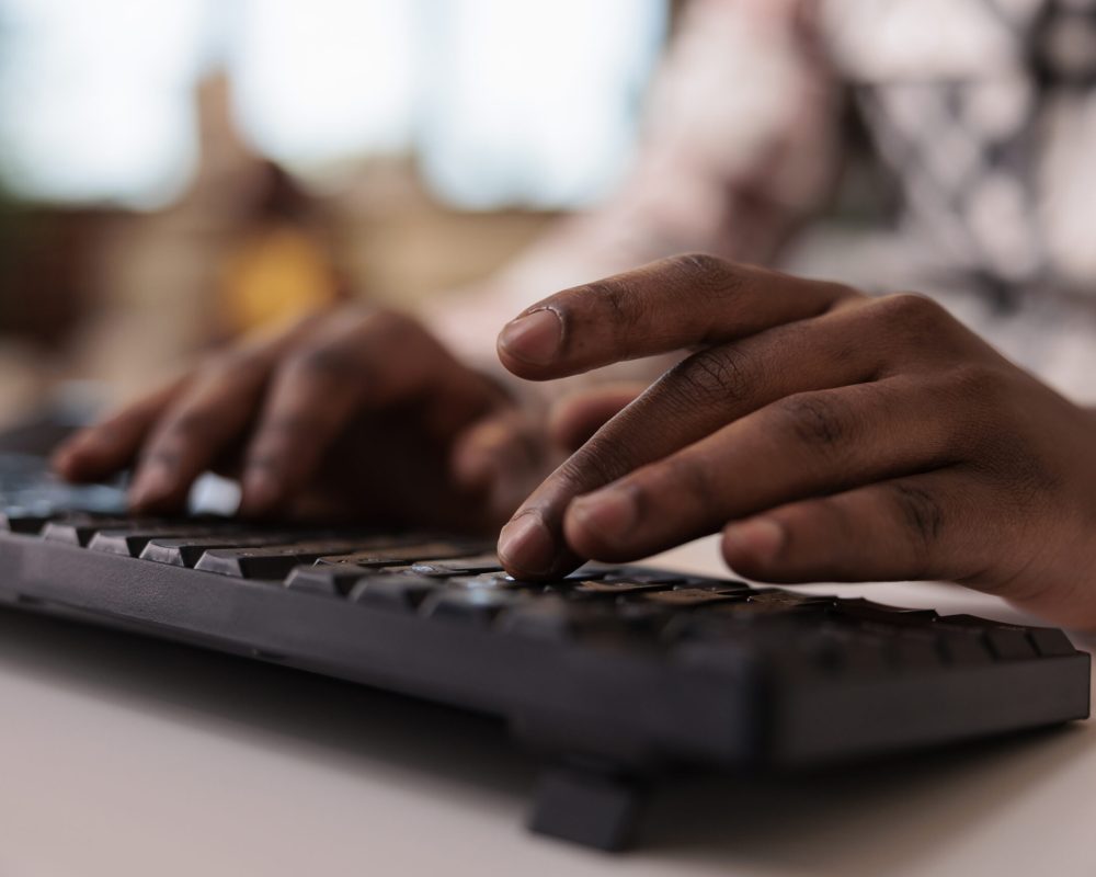 Closeup of african american male content creator writing post for social media on computer keyboard in home living room. Selective focus on man hands working remote typing blog article.